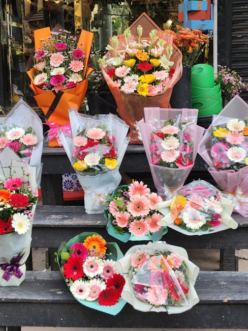 A vibrant display of assorted flower bouquets arranged on a dark wooden outdoor table, featuring a variety of fresh flowers including pink gerberas, white daisies, red and pink carnations, and yellow