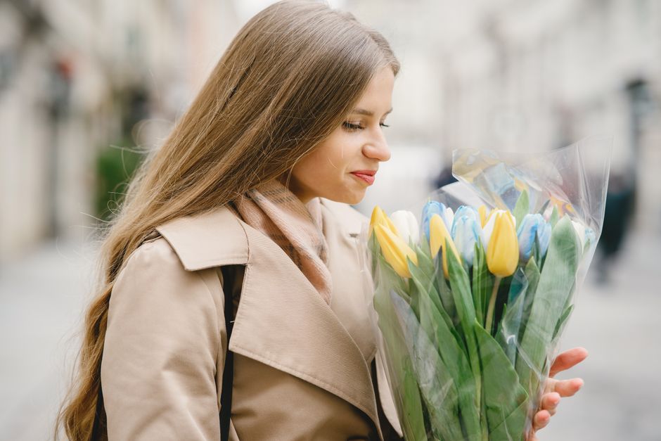 A young woman with long, light brown hair and fair skin is depicted holding a large bouquet of fresh tulips wrapped in transparent plastic. The bouquet features yellow and light blue tulips with vibra
