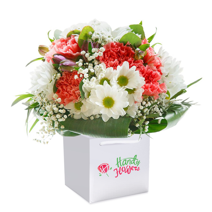 Presented upright in a sharp, modern white gift bag, this vibrant floral arrangement seems tailor-made for a Kingsbury celebration. It begins with abundant, frilled carnations in coral, peach, and deep red-their petals plush and layered, creating a soft yet spirited feel. Between these, cheerful white daisies punctuated by luminous yellow-green centers mirror the brightness and community warmth of a sunny day on Kingsbury Road. Sprigs of ethereal baby's breath are tucked throughout, lending an airy, wispy quality, while glossy, deep green leaves add structure and set the stage for the hazy pops of colour. Unopened buds flushed with purple nest along one edge, a gentle nod to new stories unfolding in the heart of Kingsbury. The skillful, rounded arrangement conveys a sense of fullness and freshness, a testament to the careful eye of the florist. The white carrier bag is both functional and elegant, with broad ribbon handles for comfortable gifting and local delivery. Set against a pure white background, the composition feels inviting and elevated, each element thoughtfully positioned to create a sense of harmony and care. Ideal for a surprise delivered on a friend's doorstep in the Kingsbury Green area, this bouquet embodies both celebration and meaningful connection within the local community.