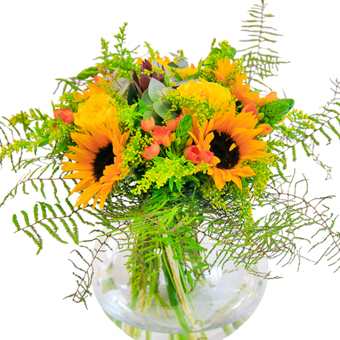 This lively arrangement, a celebration of Kingsbury's community and natural beauty, is a harmonious blend of sun-drenched colour and subtle greenery. At its center, magnificent sunflowers display their sunburst petals and velvety, coffee-toned cores, reminiscent of blooms basking in the gardens near Roe Green. Intertwined are sumptuous yellow garden roses, their layered petals opening in a slow, unfolding dance that echoes the gentle pace of mornings along Kingsbury Road. Amongst these, jewel-like red Hypericum berries glint unexpectedly, each one adding an uplifting note-much like finding moments of joy in neighbourhood gatherings. Soft fronds of delicate fern and light, ethereal sprays of goldenrod flow around the edges, drawing the eye as though tracing the undulating paths in local fields. Spikes of fresh green and silvery eucalyptus leaves complement the sunlit hues above, infusing the design with both movement and serenity. Set within a perfectly round clear glass vase, the stems and water are visible-offering a sense of refreshing clarity and craftsmanship. The bouquet's tactile interplay, from velvety petals to glossy berries to airy fern, is just as pleasing as its gentle fragrance, a subtle blend of rose and green. This arrangement, available for hand delivery throughout Kingsbury, makes a thoughtful gift to celebrate birthdays, anniversaries, or any occasion where a burst of happiness is needed.